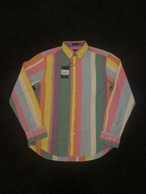 Randig skjorta från GANT i bomull - Färgglad skjorta från GANT i storlek M med breda vertikala ränder i rosa, gul, grön, orange, blå och vit. Skjortan har klassisk krage, bröstficka och knäppning framtill. Tillverkad i mjuk bomull och har lång ärm. Ny pris på 1200kr men säljs för 700 och pris kan diskuteras (Ny med prislapp)