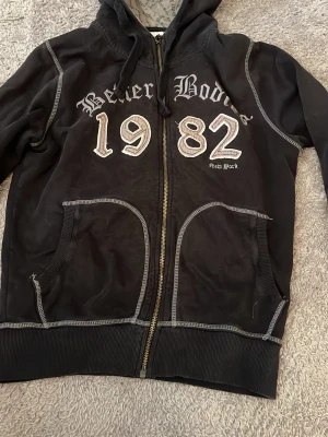 Svart hoodie Better Bodies 1982 - Svart hoodie med dragkedja från Better Bodies. Stor text '1982' och 'Better Bodies' framtill samt detaljer på ena ärmen. Gråvita kontrastsömmar och ribbade muddar. Två fickor fram och huva med snörning. Perfekt för en sportig och avslappnad stil.