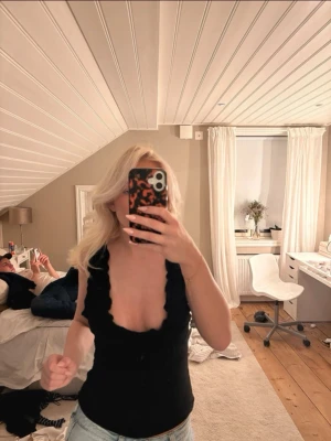 Svart topp med volangkant - Säljer en svart, tight topp med djup v-ringning och volangkant längs axelbanden och ringningen. Toppen är ärmlös och har öppen rygg, perfekt för dig som vill ha en trendig och stilren look. Passar snyggt till både jeans och kjol.
