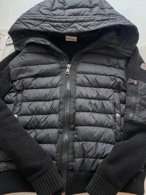 Moncler cardigan  - Snygg svart pufferjacka från Moncler med stickade ärmar och huva. Jackan har quiltad front i glansigt material, dragkedja framtill och ribbade muddar. Klassisk Moncler-logga på ärmen och praktiska fickor med tryckknappar. Perfekt för kalla dagar. Storlek 2,M men passar small 
