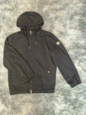 Moncler windbreaker  - Snygg svart vindjacka med huva och dragkedja framtill. Jackan har två fickor med knappstängning och ett emblem på ena ärmen. Elastiska muddar vid ärmslut och nederkant samt en rödvit rand på huvan baktill. Tillverkad i lätt, vindtätt material.