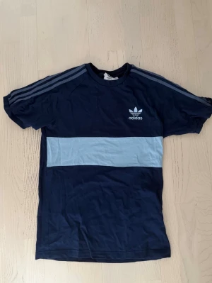 Adidas Djurgården tröja - Säljer en marinblå t-shirt från Adidas med ljusblå bred rand över bröstet och klassiska Adidas-ränder på ärmarna. T-shirten har korta ärmar och är gjord i mjuk bomull. Adidas-logga tryckt på vänster bröst. Perfekt till en sportig och avslappnad stil. Passar perfekt till DJURGÅRN MATCHER