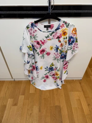 Vit blommig skjortklänning med volang - Superfin vit skjortklänning med färgglatt blommönster i rosa, blått och gult. Klänningen har volangdetaljer längs ärmarna och en lös passform. 