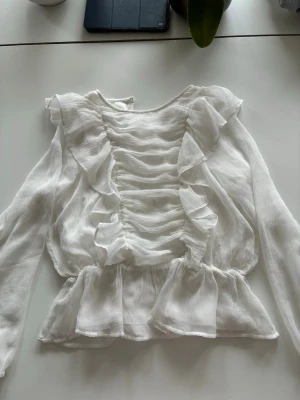 Vit söt blus  - Supersöt vit blus med volanger och rynkade detaljer framtill. Blusen har långärmad design och är gjord i ett tunt, lätt och något transparent tyg som ger en luftig känsla. Toppen är från Lindex och är i storlek 140cm.