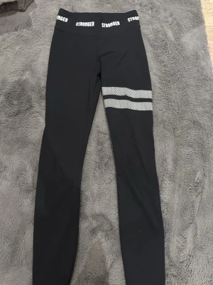 Svarta leggings från Stronger - Snygga svarta leggings från Stronger med vita loggor i midjan och två vita randiga detaljer på ena benet. Tight passform och stretchigt material som passar perfekt till träning eller chill. Midjehög modell för extra komfort.