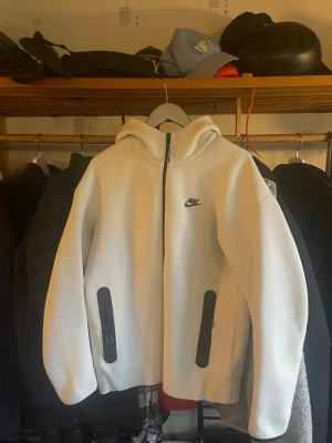 Nike teck hoddie - Säljer en stilren beige hoodie från Nike med svart dragkedja framtill och två svarta fickor med dragkedja. Hoodien har en klassisk huva och Nike-logga på bröstet. Perfekt för dig som vill ha en enkel men snygg look.