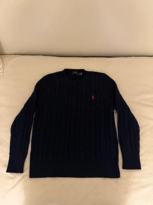 Mörkblå kabelstickad tröja Polo Ralph Lauren - Kabelstickad mörkblå tröja från Polo Ralph Lauren med rund halsringning och röd broderad logga på bröstet. Långa ärmar och ribbade muddar vid ärmslut och nederkant. Klassisk och tidlös design i mjukt material.