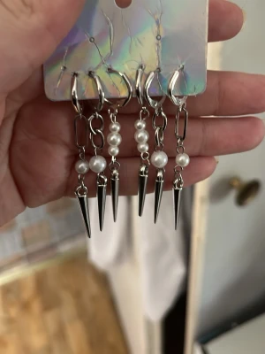 Silvriga örhängen med pärlor och spikes - Säljer fyra par coola örhängen i silverfärg med vita pärlor och spetsiga spikes. Örhängena har en modern design med kedjedetaljer och passar perfekt för dig som vill sticka ut. Materialet ser ut att vara metall och plastpärlor.