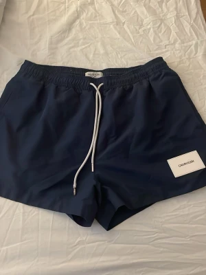 Mörkblå badbyxor Calvin Klein - Snygga mörkblå badbyxor från Calvin Klein med vit dragsko i midjan och logga på vänster ben. Klassisk modell med elastisk midja och fickor. Perfekta för stranden eller poolen.