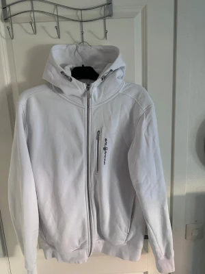 Vit hoodie från Sail Racing - Snygg vit hoodie med dragkedja från Sail Racing . Har en stor huva med dragsko och en bröstficka med dragkedja och logga. Mjuk och skön bomullsmix, perfekt för chill dagar. Klassisk passform och ribbade muddar vid ärmar och midja. Använd fåtal gånger, storlek L. Tveka inte att höra av dig för mera frågor 