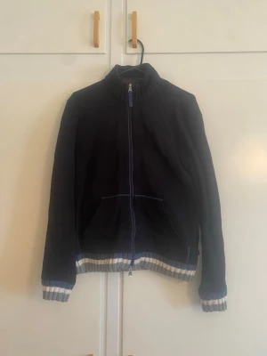 Hugo Boss svart tröja - Snygg mörkblå bomberjacka med dragkedja framtill och ribbade muddar i vitt och blått vid ärmslut och nederkant. Jackan har hög krage och två fickor framtill. Perfekt för dig som gillar en stilren och sportig look.