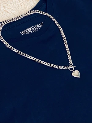 Silverfärgat halsband med hjärthänge - Snyggt halsband i silverfärg med en kedja och ett hjärthänge. Halsbandet har ett T-lås framtill som ger en cool detalj. Perfekt accessoar för att lyfta din outfit med en trendig touch. Materialet ser ut att vara rostfritt.