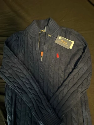 Mörkblå kabelstickad tröja Polo Ralph Lauren - Mörkblå kabelstickad tröja från Polo Ralph Lauren med halv dragkedja och hög krage. Tröjan har det klassiska röda Polo-logot på bröstet och ribbade muddar. Perfekt för dig som gillar stilren och tidlös design.