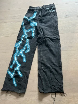 Svarta jeans med blåa blixtar - Svarta vida jeans med blått och vitt blixtmönster på ena benet och bakfickan. Knappt använda, storlek S🩵