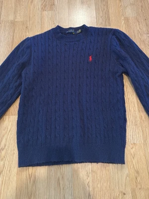Mörkblå kabelstickad tröja Polo Ralph Lauren - Kabelstickad mörkblå tröja från Polo Ralph Lauren med rund halsringning och röd broderad logga på bröstet. Tröjan har långa ärmar och ribbade muddar vid ärmslut och nederkant. Perfekt för dig som gillar klassisk och stilren look. PRISET GÅR ATT DISKUTERA!