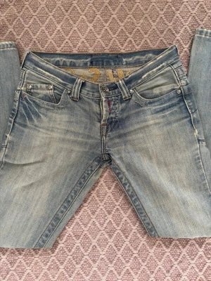 Ljusblå jeans med brodyr - Säljer ett par ljusblå jeans med slitningar och broderad text 'The Old School' i gult på baksidan. Klassisk femficksmodell med raka ben och låg midja. Materialet är denim och jeansen har coola detaljer som nitar och kontrastsömmar.