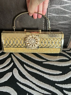 Guldig metall kuvertväska med handtag - Snygg kuvertväska i guld med glittrig yta och metallhandtag. Väskan är i metall och det finns en kedja i väskan. Små repor på väskan se bild 3 och 4 Perfekt accessoar för att lyfta din outfit med extra bling. Det fanns ingen lapp -> inget märke däremot  är den från USA/ Kanada 