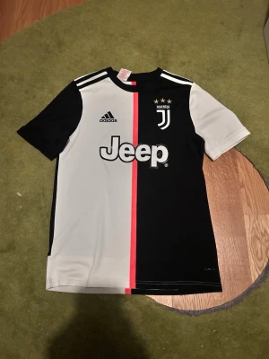 Juventus matchtröja Ronaldo 7 L i barnstorlek - Obs L i barnstorlek. Juventus officiella matchtröja från Adidas med Ronaldo 7 på ryggen. Tröjan är svart och vit med en rosa rand i mitten, Juventus-logga och Jeep som sponsor på bröstet. Korta ärmar och sportig passform, perfekt för fotbollsträning eller match. 