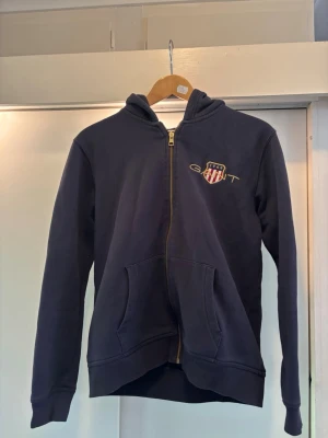 Mörkblå hoodie med dragkedja från Gant - Snygg mörkblå hoodie från Gant med dragkedja framtill och två fickor. Klassisk Gant-logga med sköld på bröstet. Huva och ribbade muddar vid ärmslut och nederkant. Perfekt för en avslappnad och stilren look.