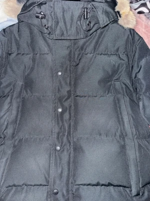 Svart pufferjacka från Canada Goose - Säljer en svart pufferjacka från Canada Goose med huva och avtagbar fuskpäls. Jackan har dragkedja och knappar framtill samt en patch på ärmen. Perfekt för kalla vinterdagar och riktigt snygg streetstil.