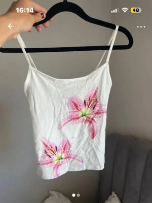 Vit linnetopp med rosa blommor - Söt vit linnetopp med tunna axelband och två stora rosa blommor framtill. Toppen har en enkel och fräsch design som passar perfekt till sommaren. Materialet känns mjukt och lätt, perfekt för varma dagar.