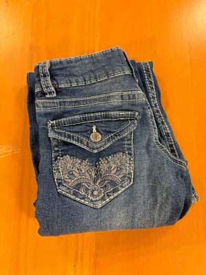 Blå bootcut jeans med broderade fickor - Säljer ett par blå bootcut jeans med snygga broderade bakfickor och dekorativa knappar. 
