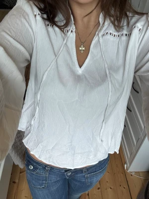 Vit blus med v-ringning - Säljer en vit blus med v-ringning och dekorativa broderade detaljer vid axlarna. Blusen har långa ärmar och en avslappnad. Perfekt att styla med jeans eller kjol för en trendig look.
