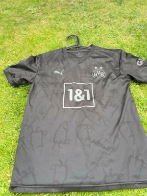 Svart Borussia Dortmund matchtröja - Säljer en svart Borussia Dortmund matchtröja från Puma med BVB-logga och två stjärnor på bröstet. Tröjan har V-ringning, korta ärmar och ett subtilt mönster i tyget. Sponsorlogga 1&1 på framsidan. Perfekt för fotbollsträning eller att bära till vardags.