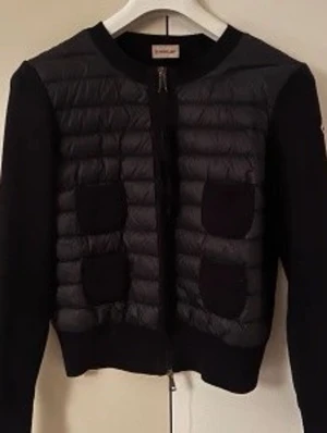 Svart quiltad cardiganjacka - Stilren svart cardiganjacka med quiltad front och fyra dekorativa fickor. Jackan har rund halsringning, dragkedja framtill och släta ärmar i kontrasterande material. Perfekt för lager-på-lager och ger en modern touch till din outfit. Taggar och nfc finns