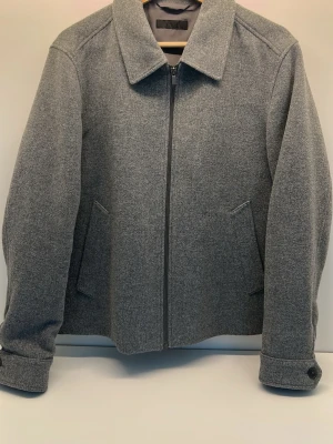 Grå ullrock med dragkedja från UNIQLO - Stilren grå rock i ullblandning med rak passform och klassisk krage. Rocken har dragkedja framtill, två snedställda fickor och knappdetaljer vid ärmsluten. Perfekt för kyliga dagar och enkel att matcha med olika outfits.