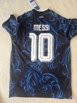 Argentina Messi tröja svart/blå - Säljer en svart och blå Argentina fotbollströja från Adidas med Messi och nummer 10 på ryggen. Tröjan har snyggt mönster i blått, vita detaljer och officiella märken på bröstet. Perfekt för dig som älskar fotboll och vill sticka ut på planen.