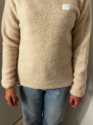 Beige teddytröja med lång ärm - Mysig beige teddytröja med lång ärm och rund hals. Tröjan har en fluffig, fårskinnsinspirerad struktur och en liten vit patch på bröstet. Perfekt för kalla dagar och enkel att matcha med jeans eller andra byxor.