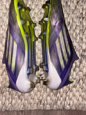 Lila och gula Adidas fotbollsskor elite f50 - Säljer ett par snabba Adidas fotbollsskor i lila och gult med vita detaljer. Skorna har en strömlinjeformad design utan snörning och tydliga dobbar för bra grepp på planen. Materialet är syntetiskt och skon har en modern look med F50-tryck på sidan.