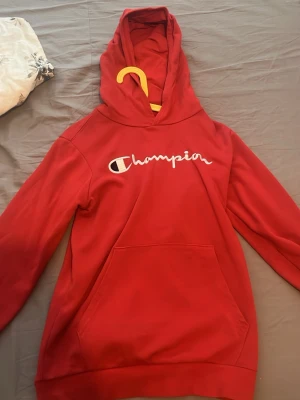Röd hoodie från Champion - Snygg röd hoodie från Champion med stor magficka och klassisk logga i vitt på bröstet. Hoodien har huva och ribbade muddar vid ärmslut och nederkant. Perfekt för en avslappnad och sportig stil.