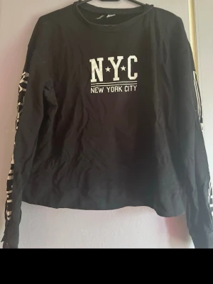 Svart långarmad tröja med NYC-tryck - Cool svart långarmad tröja med vitt NYC-tryck på bröstet och text längs ärmarna. Tröjan har rund halsringning och långa ärmar. Perfekt för dig som gillar streetstyle och vill ha en avslappnad look! Osäker på storlek med skulle tro S eller M! Trycket har mindre veck skador, som knappt syns!🌷 Spana gärna in mina andra plagg! 🤗