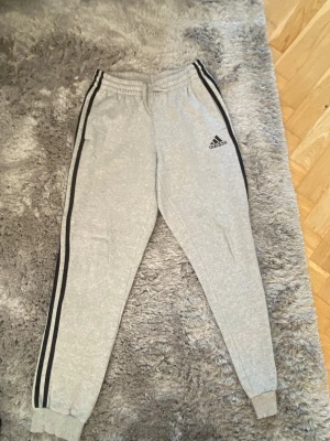 Grå Adidas mjukisbyxor med svarta ränder - Snygga grå mjukisbyxor från Adidas med klassiska svarta ränder längs sidorna och logga på vänster ben. Byxorna har resår i midjan och vid bensluten samt sidofickor. Perfekta för chill eller träning.