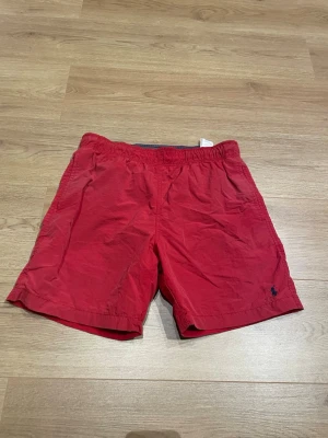 Röda badbyxor från Polo Ralph Lauren - Snygga röda badbyxor från Polo Ralph Lauren med elastisk midja och dragsko. Klassisk passform och liten broderad logga i mörkblått på ena benet. Perfekta för stranden eller poolen, tillverkade i snabbtorkande material.