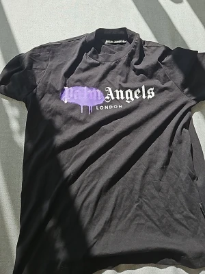Svart Palm Angels t-shirt London - Snygg svart t-shirt från Palm Angels med trycket 'Palm Angels London' på bröstet. Texten är vit med en lila drip-effekt över 'Palm'. Klassisk passform och rund hals. Perfekt för dig som gillar streetwear och vill sticka ut med en unik detalj.