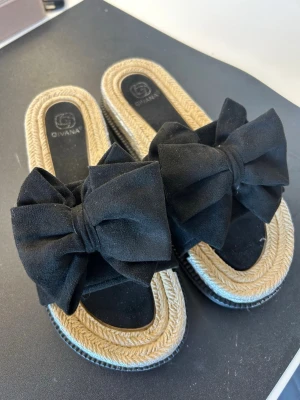 Sandaler - Snygga sandaler från Givana med stor svart rosett framtill. Ovandelen är i mjukt tyg och sulan har flätad jutedesign i beige och lite silver. De är endast använda en gång, alltså som nya. De är i storlek 40 men passar mig som vanligtvis har 39.