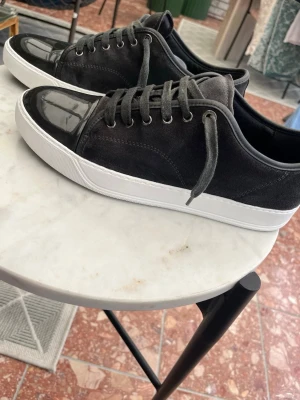 Svarta sneakers med lackdetalj - Snygga svarta sneakers med ovandel i mocka och glansig lackdetalj på tån. Skorna har vita, tjocka sulor och svarta skosnören. Perfekta för dig som vill ha en stilren men ändå unik look.