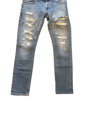 Nudie jeans  - Säljer ett par ljusblå jeans med raka ben och flera slitningar framtill för en trendig look. Jeansen har klassisk femficksdesign och normal passform. Perfekta för dig som gillar en avslappnad och edgy stil.