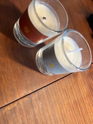 Två doftljus i glas - Säljer två små doftljus i genomskinliga glasbehållare. Ett har en brun etikett och det andra en grå, båda med vita vaxljus och citrondetaljer på etiketten. Perfekta för att skapa mysig stämning hemma.