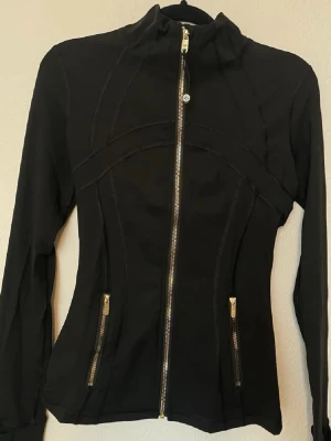 Lululemon zip up Svart jacka med gulddragkedjor - Snygg svart jacka med figurnära passform och guldfärgade dragkedjor framtill och på fickorna. Jackan har hög krage och långa ärmar, perfekt för en sportig och stilren look. Materialet känns mjukt och stretchigt.