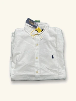 Vit linneskjorta från Polo Ralph Lauren - Klassisk vit skjorta i linne från Polo Ralph Lauren med button-down krage och broderad marinblå logga på bröstet. Skjortan har långa ärmar och knappar framtill. Perfekt för en clean och stilren look.