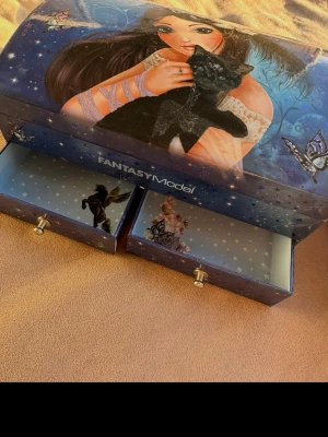 Fantasy Model smyckeskrin med lådor - Blått smyckeskrin från Fantasy Model med två utdragbara lådor. Locket har en färgglad illustration av en tjej med långt hår och en svart katt, samt fjärilar och stjärnor. Insidan av lådorna är dekorerade med motiv av älvor och stjärnor. Perfekt för att förvara smycken och småaccessoarer. 🌷 Spana även in andra saker jag säljer!🤗