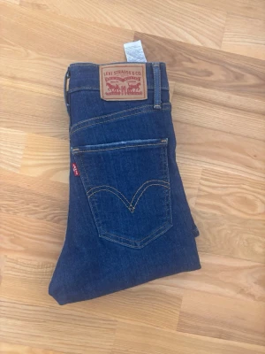 Levi's blå skinny jeans - Säljer ett par klassiska blå skinny jeans från Levi's med hög midja och fem fickor. Jeansen har den ikoniska röda Levi's-lappen på bakfickan och kontrastsömmar. Perfekta för en avslappnad och trendig look.
