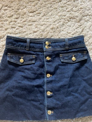 Mörkblå jeanskjol med knappar H&M - Snygg mörkblå jeanskjol från H&M med guldiga knappar framtill och två dekorativa fickor. Använd vid 2 tillfällen.