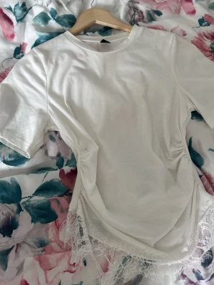 Vit topp med spetsdetalj och rynk - Säljer en vit topp med korta ärmar, rynkade sidor och spetsdetalj längst ner. Toppen har en rund halsringning och är gjord i mjukt material som känns skönt mot huden. Perfekt för dig som gillar en feminin och trendig stil.