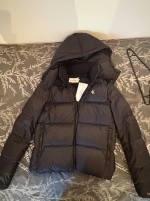 Svart pufferjacka från Calvin Klein - Snygg svart pufferjacka från Calvin Klein Jeans med huva och dragkedja framtill. Jackan har två stora fickor med dragkedja och diskret CK-logga på bröstet. Perfekt för kalla vinterdagar och riktigt skön att bära. Materialet är glansigt och quiltat. Köpt från JeansBolaget och har även kvar kvittot. Pris kan diskuteras ner. Helt ny med lappar kvar.