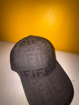 Svart keps med Fendi-mönster - Snygg svart keps från Fendi med det klassiska FF-mönstret över hela kepsen. Kepsen har böjd skärm och är tillverkad i ett tygmaterial som ger en exklusiv känsla. Perfekt accessoar för dig som vill sticka ut med stil.